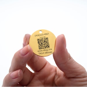 Op de afbeelding: Een ronde metalen tag in goudkleur met een zwart-witte QR-code, de tekst "SCAN ME:" en de tekst "www.idtag.link TAG#:dBzoa0" en het telefoonnummer (416) 123-4567.