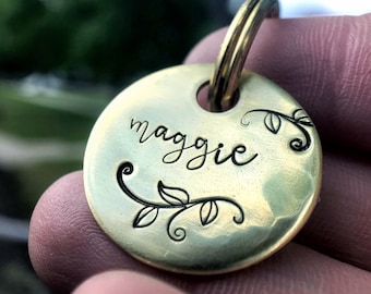 May include: A circular brass pet tag engraved with the name "maggie" in cursive. The tag is adorned with a leaf and vine pattern and includes a brass ring for attachment.
