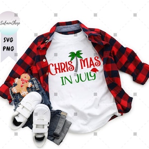 May include: A white t-shirt with red and green text that says "Christmas in July" with a palm tree and Santa hat graphic. The shirt is folded and displayed on a red and black plaid shirt and blue jeans.