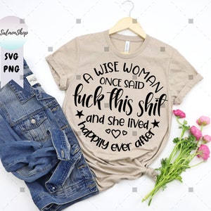 May include: Beige t-shirt with the text "A WISE WOMAN ONCE SAID fuck this shit and she lived happily ever after" in a black, circular design. A denim jacket and flowers are next to the shirt. The shirt is hanging on a wooden hanger.