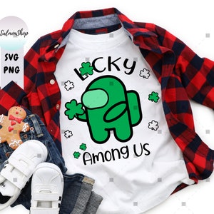 Puede incluir: Una camiseta blanca con un personaje verde de "Among Us" sosteniendo un tr&eacute;bol de cuatro hojas. El texto "Lucky" est&aacute; por encima del personaje y "Among Us" por debajo. La camiseta est&aacute; sobre una camisa de cuadros rojos y negros.