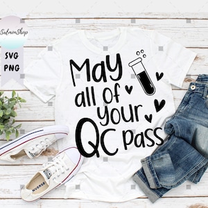 May include: White t-shirt with black text that reads "May all of your QC pass" with a test tube and hearts. The shirt is laid out on a woodgrain surface with a pair of white sneakers and blue jeans.