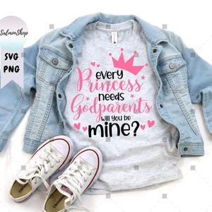 May include: A light gray t-shirt with the text "every princess needs Godparents will you be mine?" in pink and black, with a pink crown and hearts. The shirt is paired with a light blue denim jacket and white sneakers.