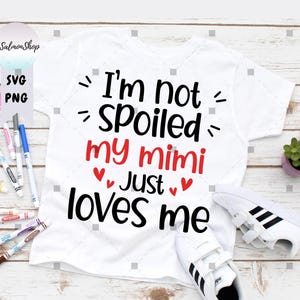 May include: White t-shirt with the text "I'm not spoiled my mimi just loves me" in black and red. The shirt is paired with white trainers with black stripes. Markers and a small plant are also visible.