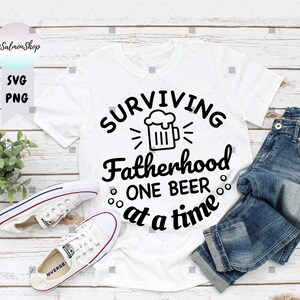 May include: White t-shirt with black text that reads "Surviving Fatherhood One Beer at a Time". A beer mug graphic is above the text. The shirt is laid out on a woodgrain surface with a pair of white sneakers and blue jeans.