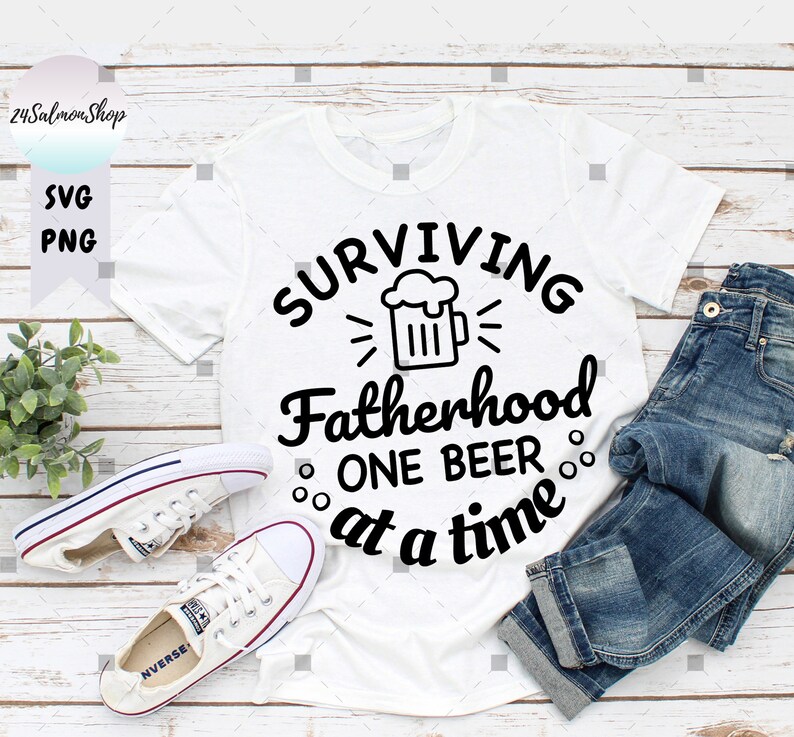 May include: White t-shirt with black text that reads "Surviving Fatherhood One Beer at a Time". A beer mug graphic is above the text. The shirt is laid out on a woodgrain surface with a pair of white sneakers and blue jeans.
