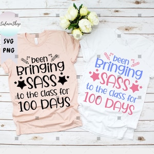 Może przedstawiać: Dwie koszulki z nadrukiem "been bringing sass to the class for 100 days". Jedna koszulka jest r&oacute;żowa, a druga biała.