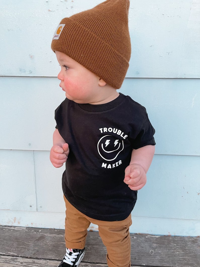 May include: A toddler wearing a black t-shirt with a white smiley face graphic that says "Trouble Maker" and a brown beanie. The toddler is wearing brown pants and black shoes.