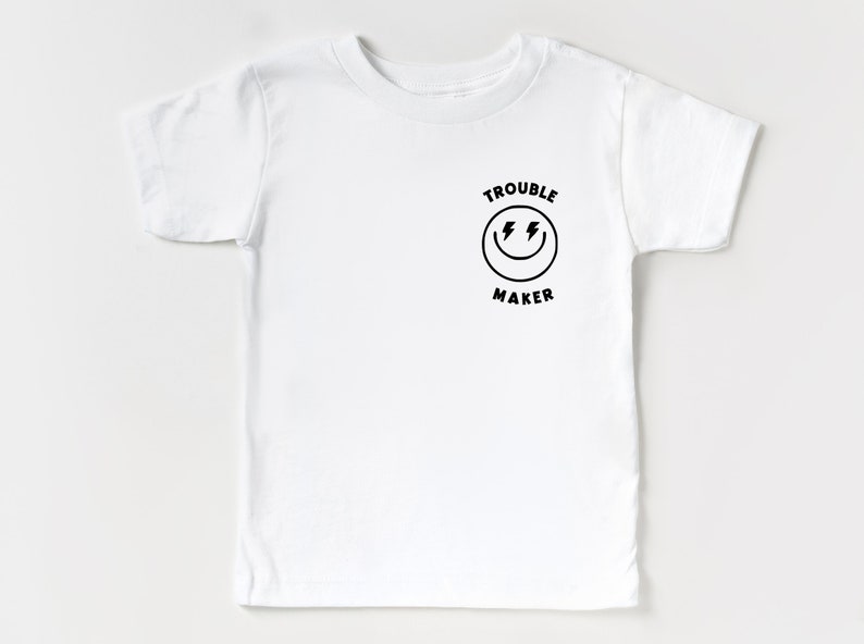 May include: White short-sleeved t-shirt with a black graphic of a smiley face with lightning bolts for eyes and the text "Trouble Maker" above and below the face.