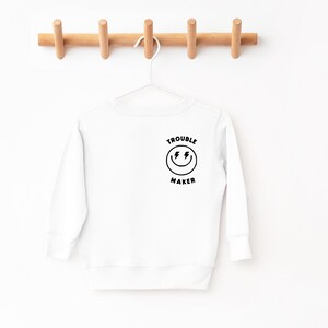 May include: White long-sleeve sweatshirt with a black smiley face graphic and the text "Trouble Maker" on the back.
