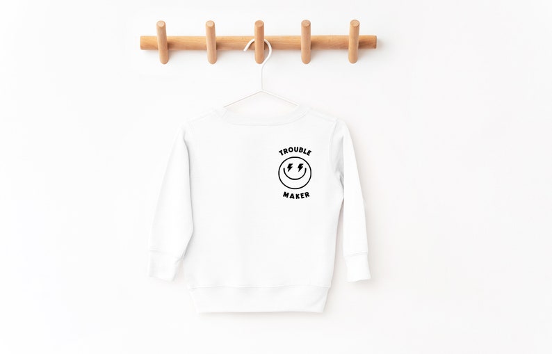 May include: White long-sleeve sweatshirt with a black smiley face graphic and the text "Trouble Maker" on the back.