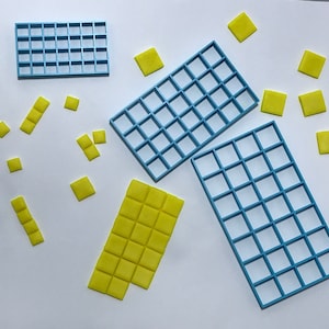 Puede incluir: Tres moldes de rejilla cuadrada de plástico azul para hacer cuadrados de fondant o chocolate. También hay muchos cuadrados de fondant amarillos esparcidos alrededor de los moldes.