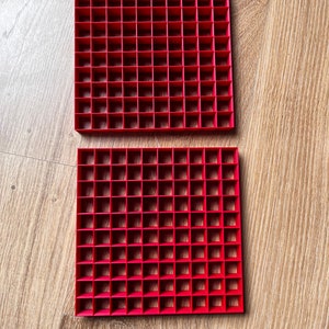 May include: Two red plastic grid trays with square compartments. The trays are stacked on top of each other.