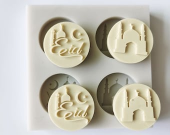 Puede incluir: Un molde de silicona gris claro con cuatro cavidades circulares. Dos cavidades presentan la palabra "Eid" en escritura con una mezquita y una luna creciente. Las otras dos tienen un diseño de mezquita. Para jabones o velas.