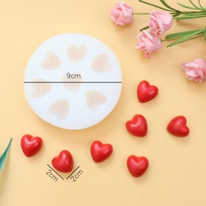 May include: A white silicone mold with six heart-shaped cavities, measuring 9 cm in diameter.  The mold is surrounded by seven small red heart-shaped candies, each measuring 2 cm in diameter.