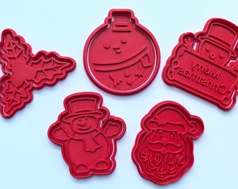 May include: Five red plastic Christmas cookie cutters. The cutters are shaped like a holly leaf, a snowman, a Santa Claus, a Christmas ornament, and a sign that says "Merry Christmas".