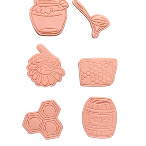 Könnte beinhalten: Sechs rosa Plastik-Ausstecher in Form von Honig-Motiven: ein Glas, ein Löffel, eine Wabe, eine Blume, eine Wabe und ein Fass mit der Aufschrift "Honey".