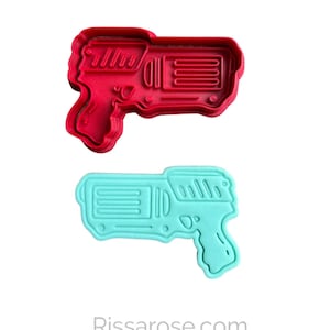 Puede incluir: Dos cortadores de galletas de plástico con forma de pistolas de agua. Uno es rojo y el otro es azul claro.