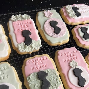 Może przedstawiać: Zbliżenie na sześć ozdobionych ciasteczek cukrowych z różowym i zielonym lukrem. Ciasteczka są ozdobione tekstem "FATIMAS TEA PARTY" i sylwetką kobiety. Ciasteczka znajdują się na czarnym drucianym stojaku.
