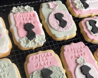 May include: A close-up of six decorated sugar cookies with pink and green icing. The cookies are decorated with the text "FATIMAS TEA PARTY" and a silhouette of a woman. The cookies are on a black wire rack.