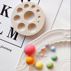 Può includere: Uno stampo in silicone beige con incavi circolari di varie dimensioni. Caramelle rotonde multicolori rosa, arancioni, gialle, verdi, blu e viola sono disposte su un piatto bianco. La parola "journey" è visibile sullo sfondo.