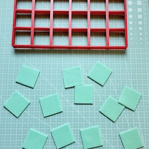 May include: A red plastic grid with 25 squares, and 13 turquoise squares of various sizes on a green cutting mat.