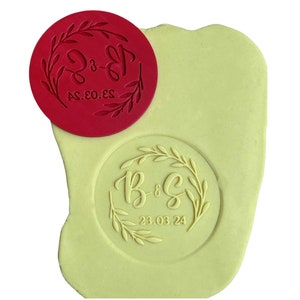 May include: A red rubber stamp with a wreath design and the text "B&S 23.03.24" in a white font. The stamp is on top of a yellow clay surface with an impression of the stamp.