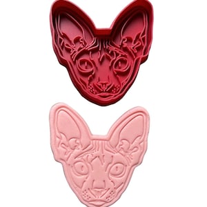 Puede incluir: Un cortador de galletas de plástico rojo y rosa con forma de cabeza de gato Sphynx. El cortador está detallado con los rasgos faciales del gato, incluidos sus ojos, nariz y bigotes.
