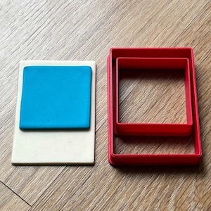 Peut inclure: Deux emporte-pièces carrés en plastique rouge, l'un à l'intérieur de l'autre, sur une surface en bois. Un carré bleu de pâte à modeler est sur un carré de papier blanc.