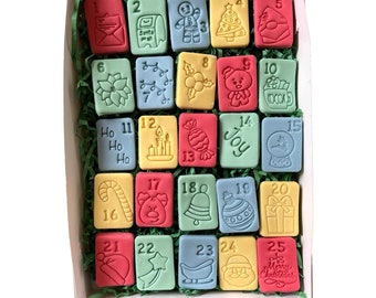 Peut inclure: Un calendrier de l'avent de Noël avec 25 cases individuelles, chacune avec un motif festif différent et un numéro. Les cases sont disposées en grille de 5x5 et sont faites d'un matériau ressemblant à du fondant coloré. Le calendrier est dans une boîte blanche avec du papier déchiqueté vert.