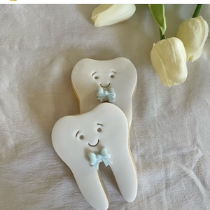 May include: Two white sugar cookies decorated to look like teeth with a blue bow on each. The cookies are on a white surface.