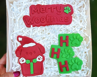Puede incluir: Una caja de regalo roja y verde con temática navideña con un perro que lleva un gorro de Papá Noel y sostiene un regalo. La caja también incluye un letrero rojo que dice "Merry Woofmas" y un letrero verde que dice "Ho Ho Ho" con huellas de patas.