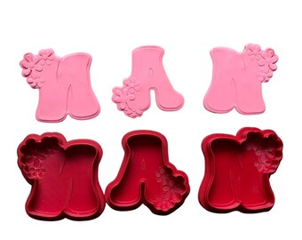 May include: Two sets of pink and red plastic cookie cutters shaped like the letters "M" and "A" with floral designs. The cutters are for making cookies or other baked goods.