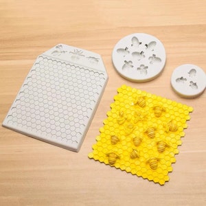 Pode incluir: Três moldes de silicone para assar, com um padrão de favo de mel e desenhos de abelhas. Os moldes são brancos e o padrão de favo de mel é cinza. Os desenhos de abelhas estão em um círculo e um retângulo. O padrão de favo de mel também é mostrado em um molde de silicone amarelo com desenhos de abelhas.
