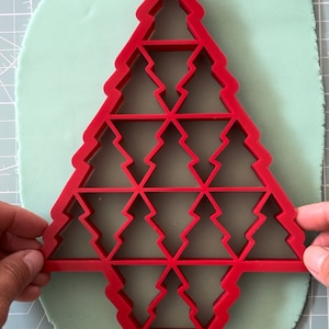 May include: Red plastic Christmas tree cookie cutter with a design of multiple smaller trees inside the larger tree shape.