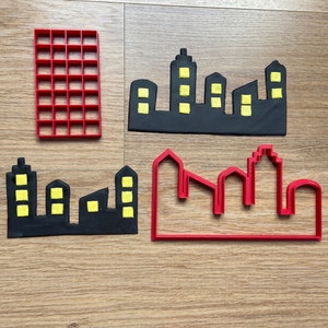 Può includere: Quattro tagliabiscotti in plastica rossa a forma di skyline di città. Due tagliabiscotti sono pieni e due sono contornati. I tagliabiscotti pieni hanno finestre gialle.