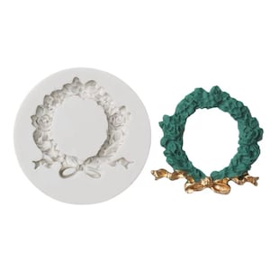 May include: A white silicone mould for making a green floral wreath with a gold bow. The mould is shaped like a wreath with a bow at the bottom. The wreath is decorated with flowers and leaves.