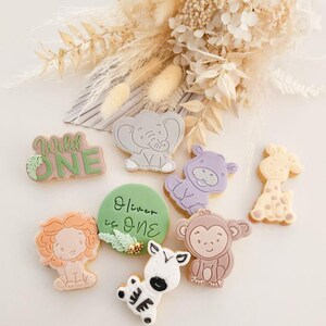 May include: A set of nine decorated cookies featuring a variety of jungle animals, including a lion, zebra, giraffe, monkey, elephant, and hippopotamus. The cookies are decorated with colourful icing and have a whimsical, playful design. One cookie reads "Wild One" and another reads "Oliver is One".