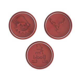 May include: Three red circular cookie cutters with white borders. One features an elf face, another a reindeer head, and the third has the text "I'm Back!" with elf feet. Festive tools for holiday baking.