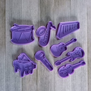 Pode incluir: Um conjunto de nove cortadores de biscoitos de plástico roxo em forma de instrumentos musicais, incluindo um tambor, um saxofone, um teclado, um violão, uma guitarra elétrica, um violino, um microfone e um piano.