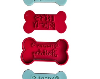 May include: Four dog bone-shaped cookie cutters. Two are red and two are light blue. The red cutters have the text "Let's Paw-ty" and "Happy Birthday" in white. The blue cutters have the text "Let's Paw-ty" and "Happy Birthday" in green.