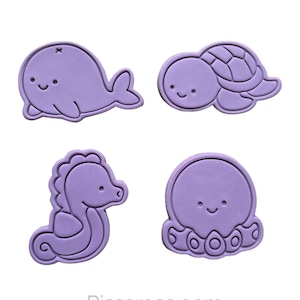 Może przedstawiać: Zestaw czterech fioletowych plastikowych foremek do ciasteczek w kształcie wieloryba, żółwia, konika morskiego i ośmiornicy. Formy są zaprojektowane z prostymi, kreskówkowymi twarzami.