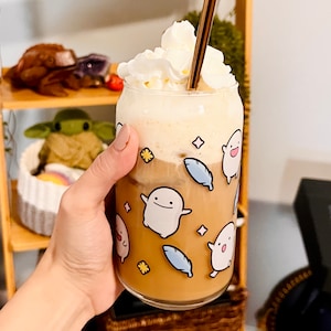 Pu&ograve; includere: Un bicchiere di caff&egrave; trasparente pieno di una bevanda marrone chiaro, sormontato da panna montata e una cannuccia dorata. La tazza &egrave; decorata con personaggi di fantasmi dei cartoni animati, piume blu e forme di stelle e fiori.