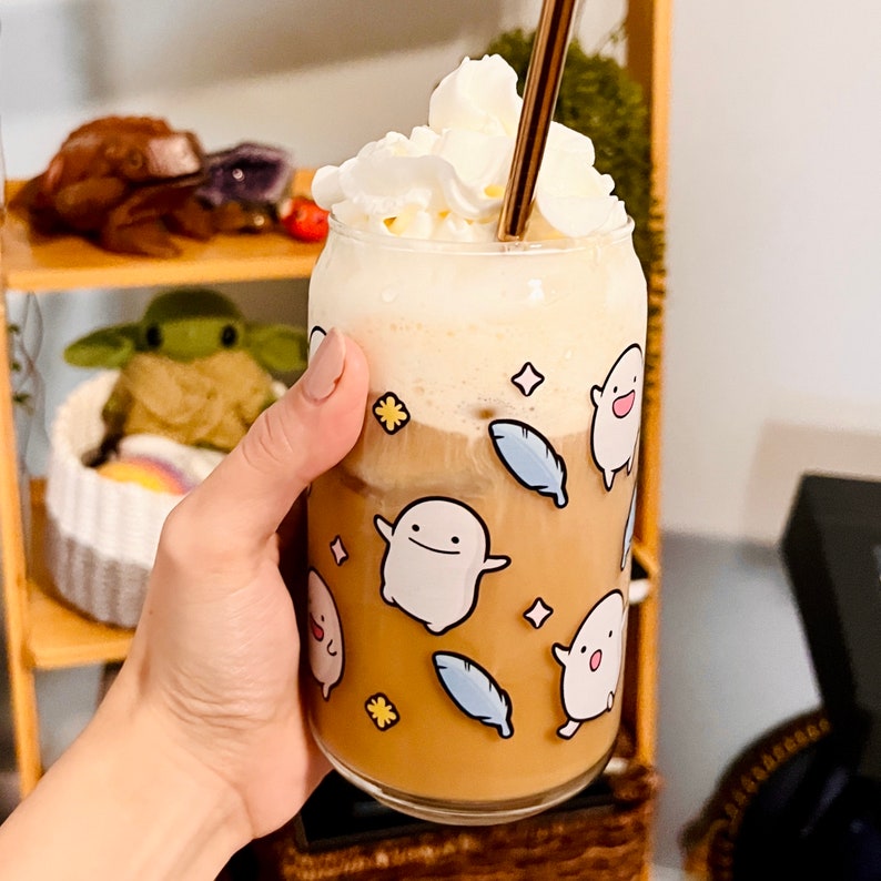 Pu&ograve; includere: Un bicchiere di caff&egrave; trasparente pieno di una bevanda marrone chiaro, sormontato da panna montata e una cannuccia dorata. La tazza &egrave; decorata con personaggi di fantasmi dei cartoni animati, piume blu e forme di stelle e fiori.