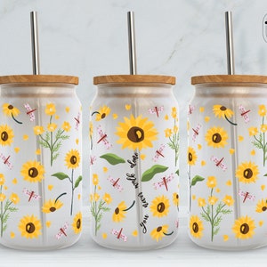 May include: Three clear glass tumblers with lids and straws. Each tumbler has a white background with a pattern of yellow sunflowers, green leaves, pink dragonflies, and small white flowers. The middle tumbler has the text "You never walk alone" written vertically on the sunflower.