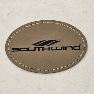 May include: A brown leather oval-shaped coaster with white stitching around the edge. The coaster has a black logo that says "Southwind" with a stylized wave design.