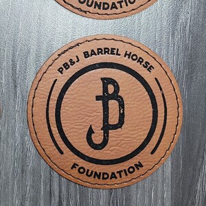 May include: Four brown leather coasters with black stitching and a black logo. The logo is a stylised letter "B" with a fish hook inside the letter. The text "PB&J BARREL HORSE FOUNDATION" is printed around the logo.