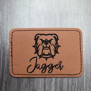 May include: A rectangular, tan leather patch with a black stitched border. The patch features a black illustration of a bulldog's head and the name "Jagger" in a cursive font.