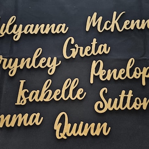 May include: A collection of gold-colored, script-style cutouts of the names Lilyanna, McKenna, Brynley, Greta, Penelope, Isabelle, Sutton, Emma, and Quinn. Each name is crafted in a cursive font and features a smooth, metallic gold finish.