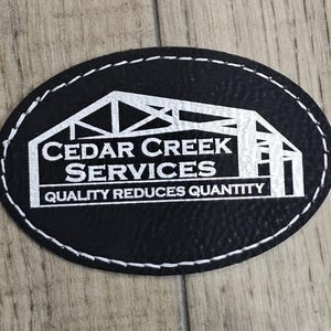 May include: An oval black patch with a textured surface and white stitched edges. The patch features a white graphic of a building frame and the text CEDAR CREEK SERVICES and QUALITY REDUCES QUANTITY printed in a bold white sans-serif font.